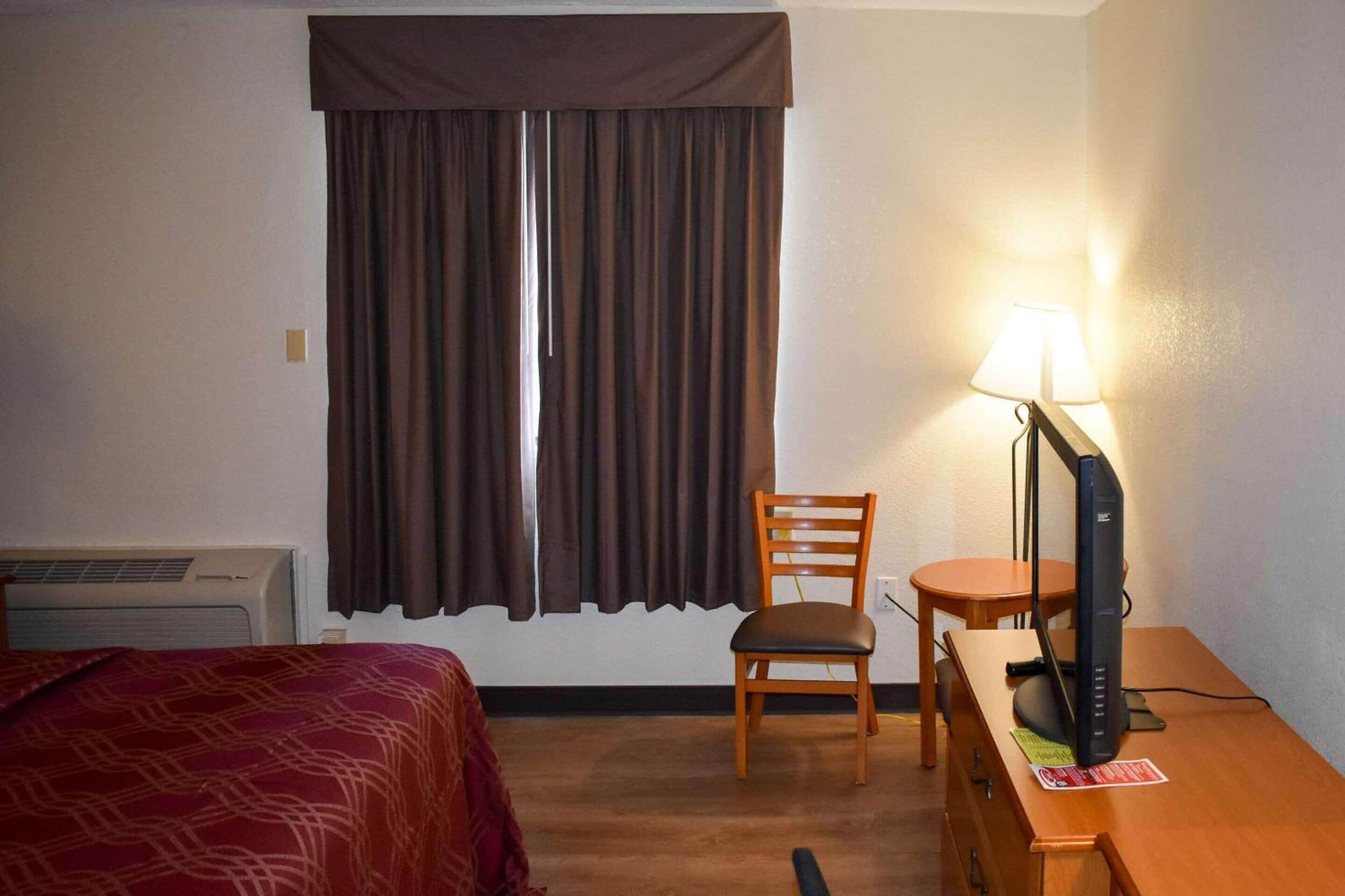 A hotel room with a bed, a desk with a computer, a chair, a lamp, and a window with curtains.