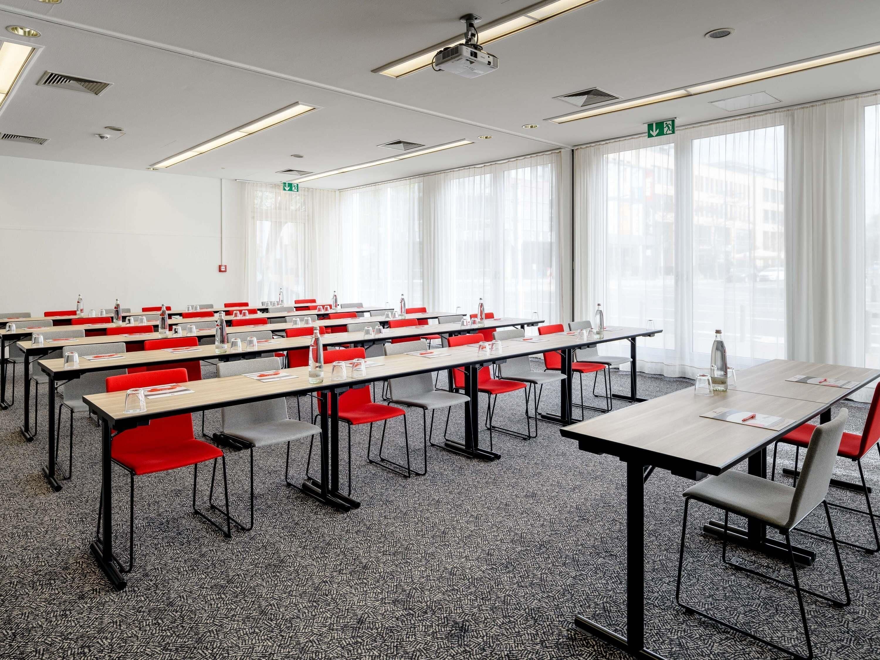Ein Konferenzraum mit mehreren Tischen und Stühlen, großen Fenstern mit Vorhängen und einem Projektor an der Decke.