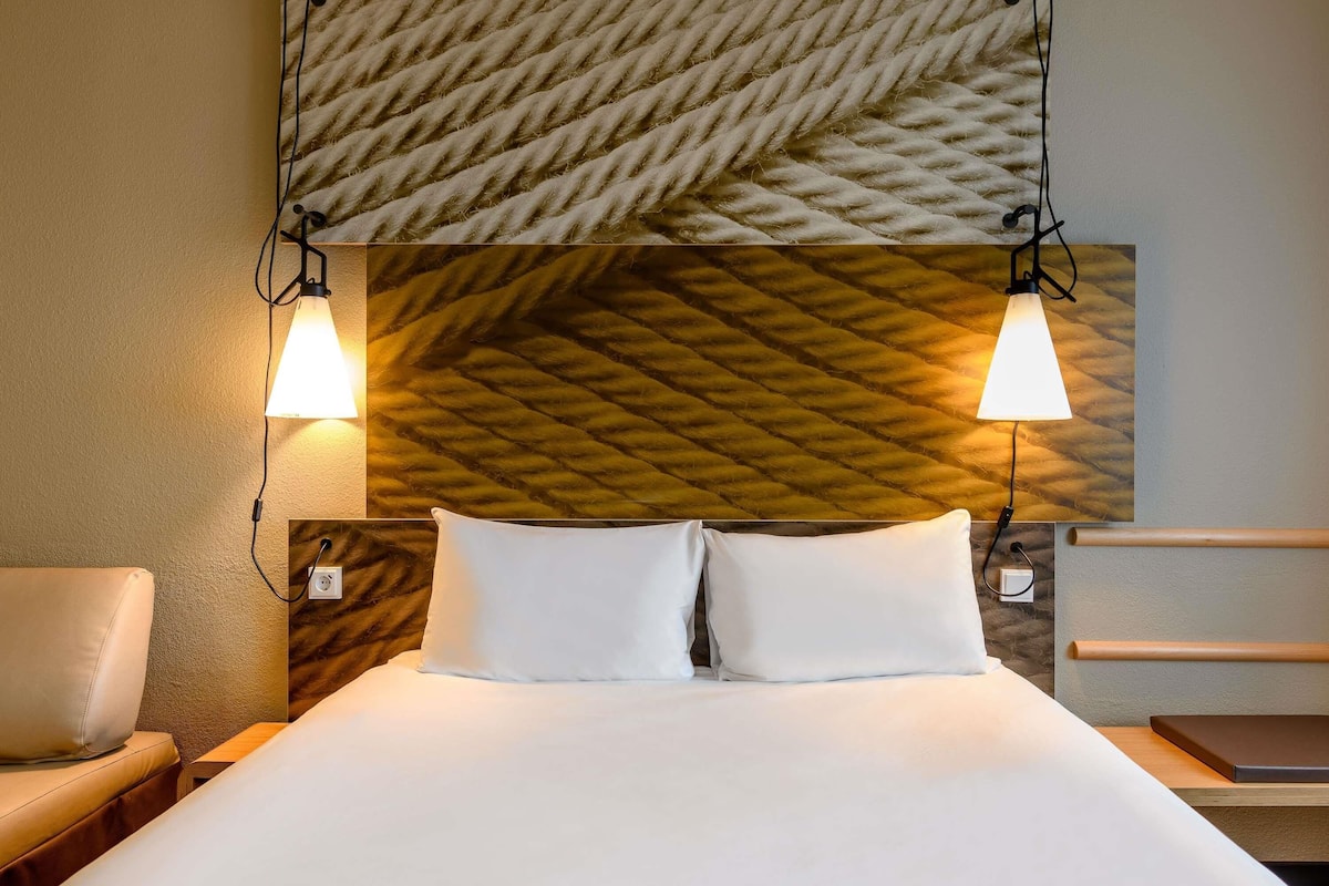 A hotel room with a bed, two bedside lamps, a chair, and a wooden headboard.