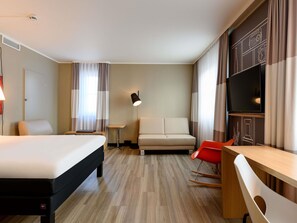 A hotel room with a bed, a sofa, a desk, a chair, a TV, and a lamp. - ibis Mainz City (Mainz)