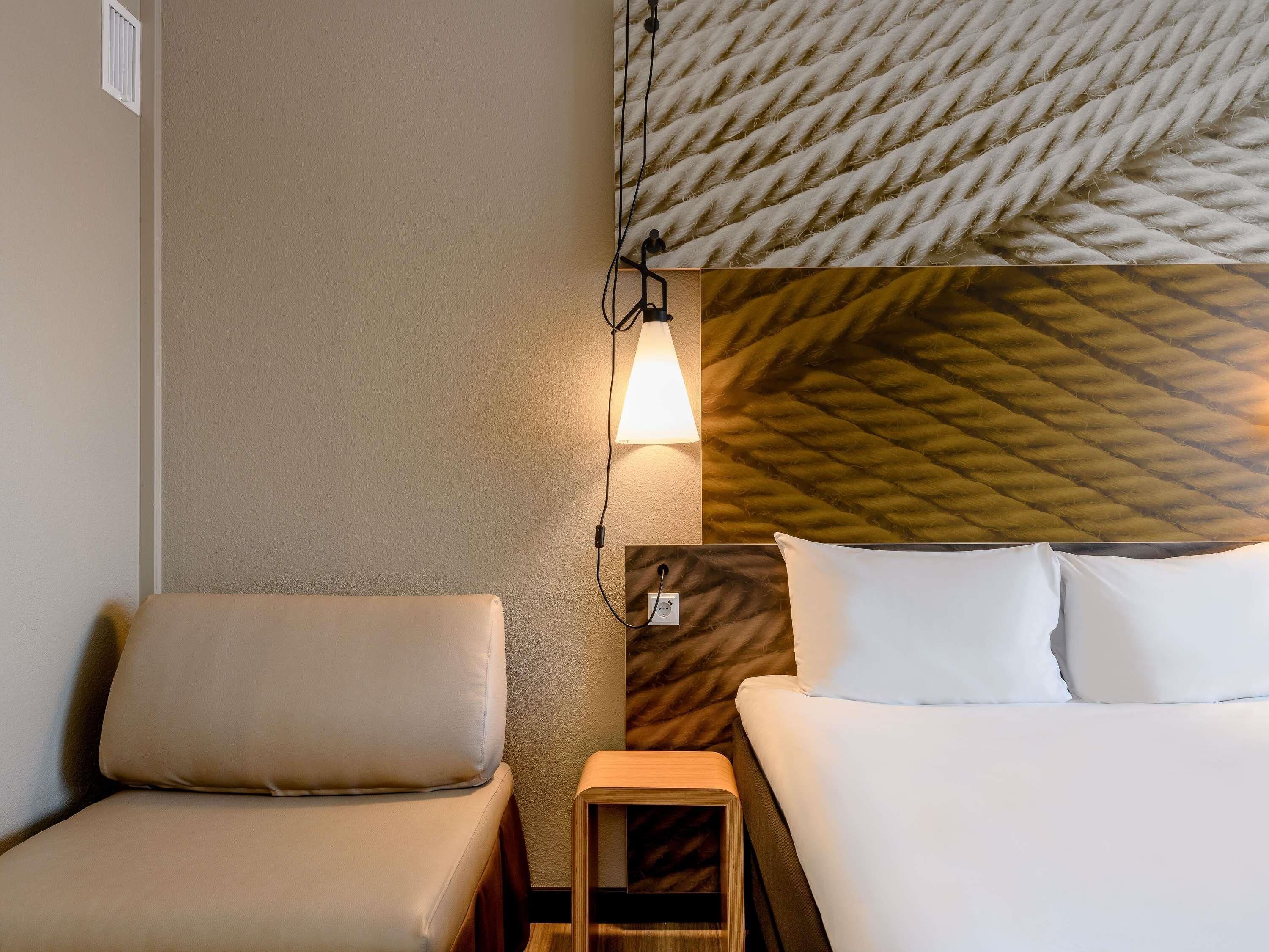 A hotel room with a bed, a chair, a nightstand, and a wall-mounted lamp.