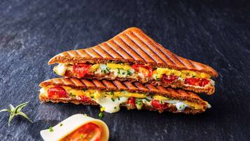 Un sándwich a la plancha con ingredientes visibles como tomates, queso y huevos, sobre una superficie oscura.