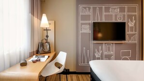 Habitación de hotel con cama, escritorio con lámpara, silla, televisor montado en la pared y ventana con cortinas.