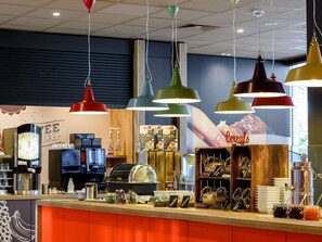A breakfast bar with various food items and condiments, overhead hanging lights, and a coffee machine. - ibis Mainz City (Mainz)