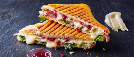 Un sándwich a la plancha cortado por la mitad, sobre una superficie oscura, con un pequeño bol de salsa roja y algunas hojas verdes cerca.