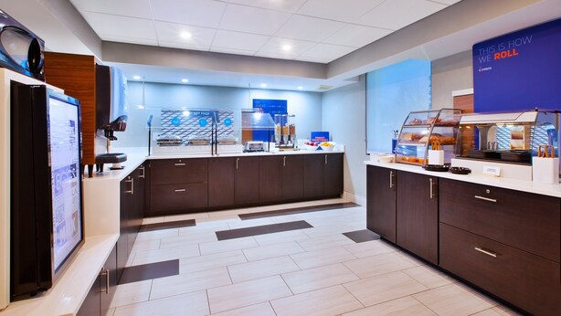A modern hotel kitchen with a buffet area, a coffee station, and a digital screen displaying information. - Holiday Inn Express Fairfax - Arlington Boulevard by IHG (Fairfax)