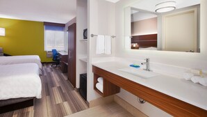 Habitación de hotel con una cama grande, un escritorio, una silla, baño con lavamanos y espejo, y una ventana con persianas.