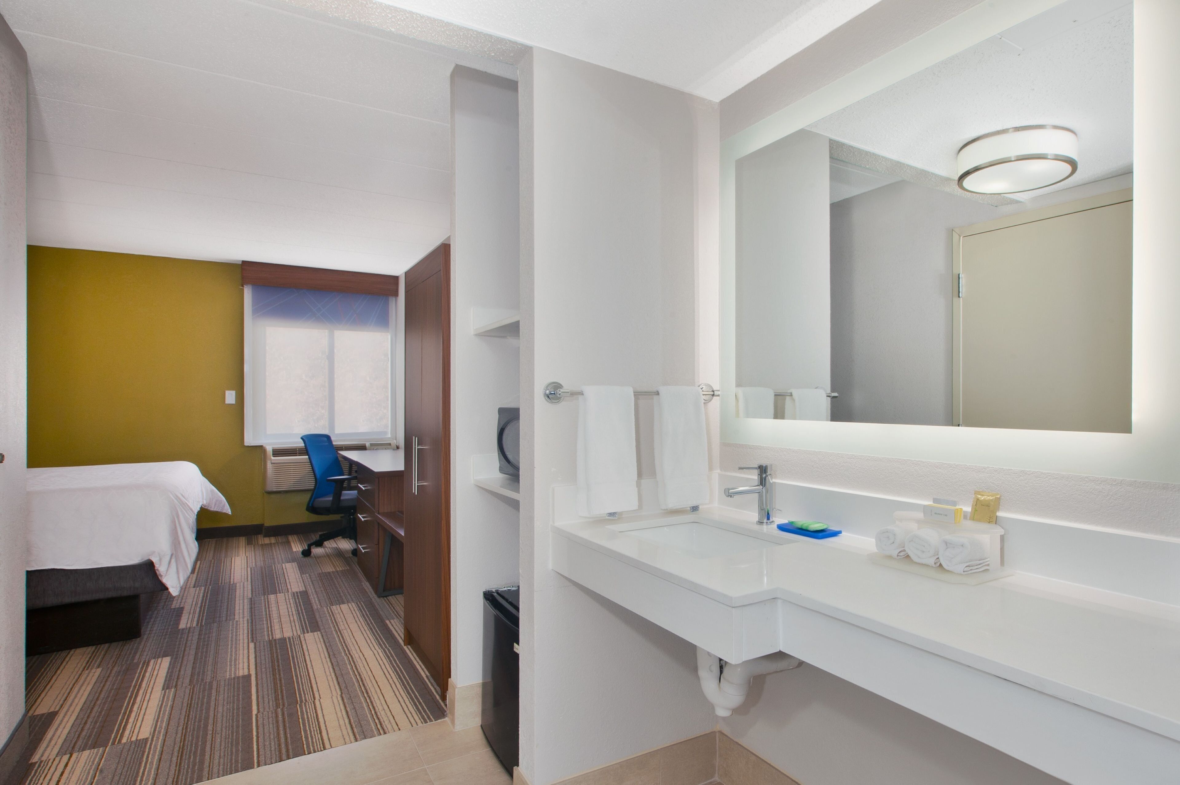 Una habitación de hotel con cama, escritorio, silla y baño con lavamanos y espejo.