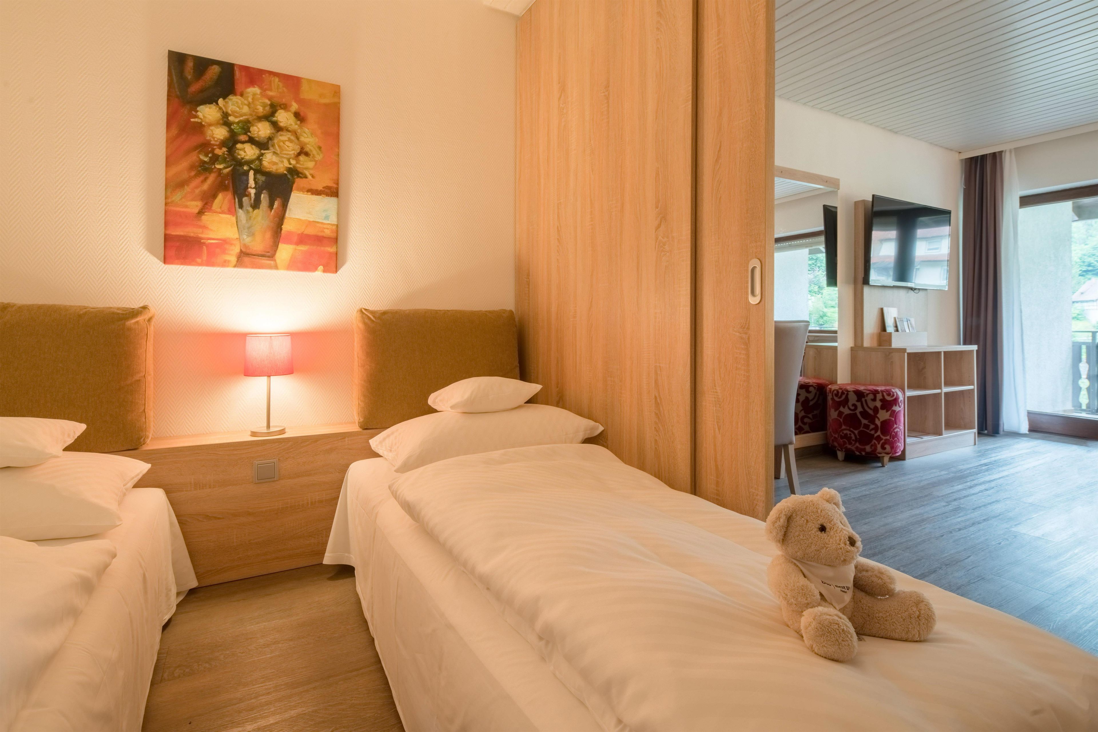A hotel room with two beds, a painting, a lamp, a teddy bear, and a view of the outside.