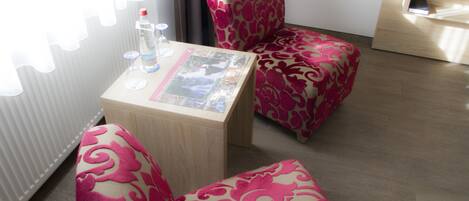 Um quarto pequeno e moderno com piso de madeira, uma mesa de vidro com uma revista e duas cadeiras estampadas.
