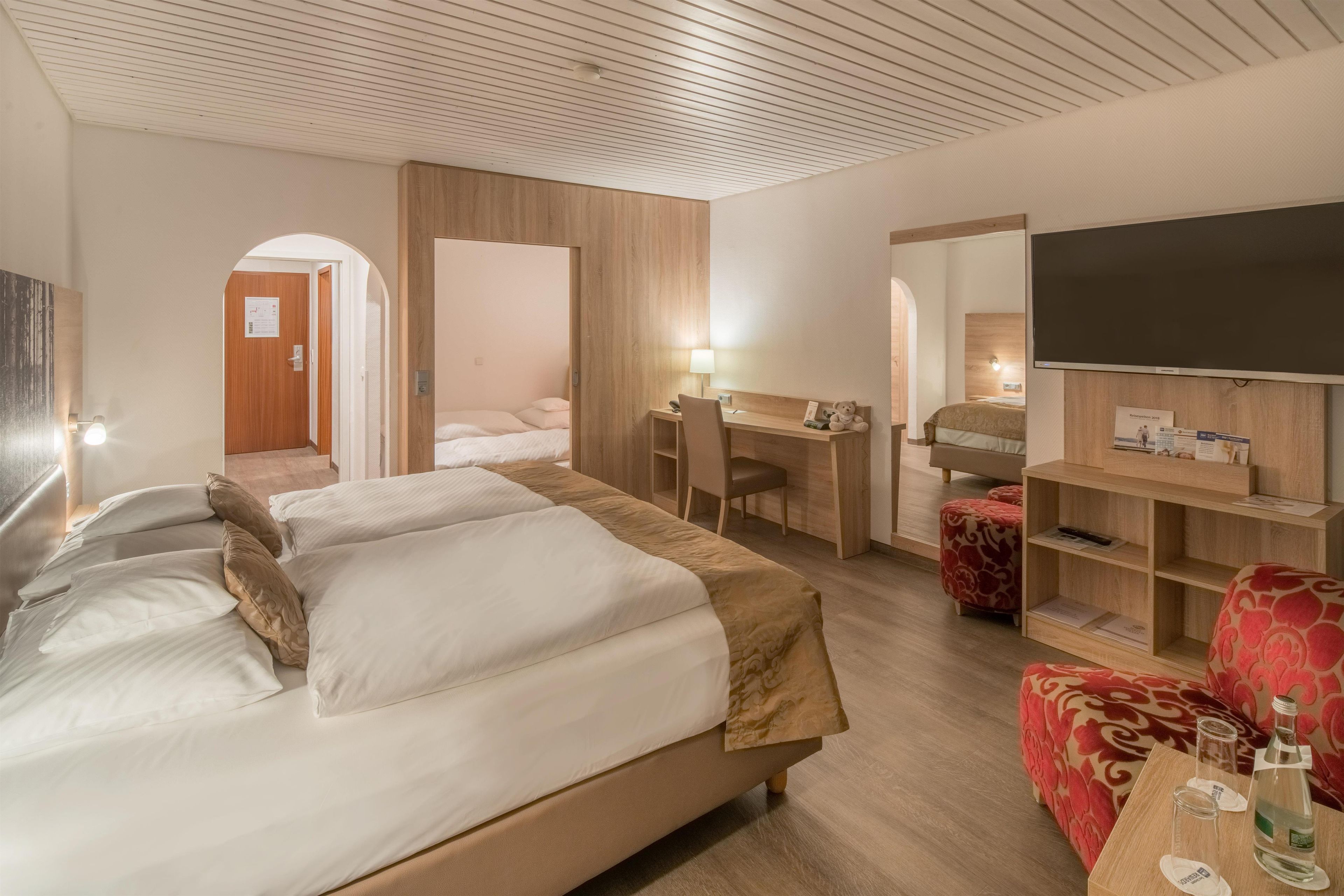 A hotel room with a large bed, a desk, a chair, a TV, and a small table with glasses and a bottle.