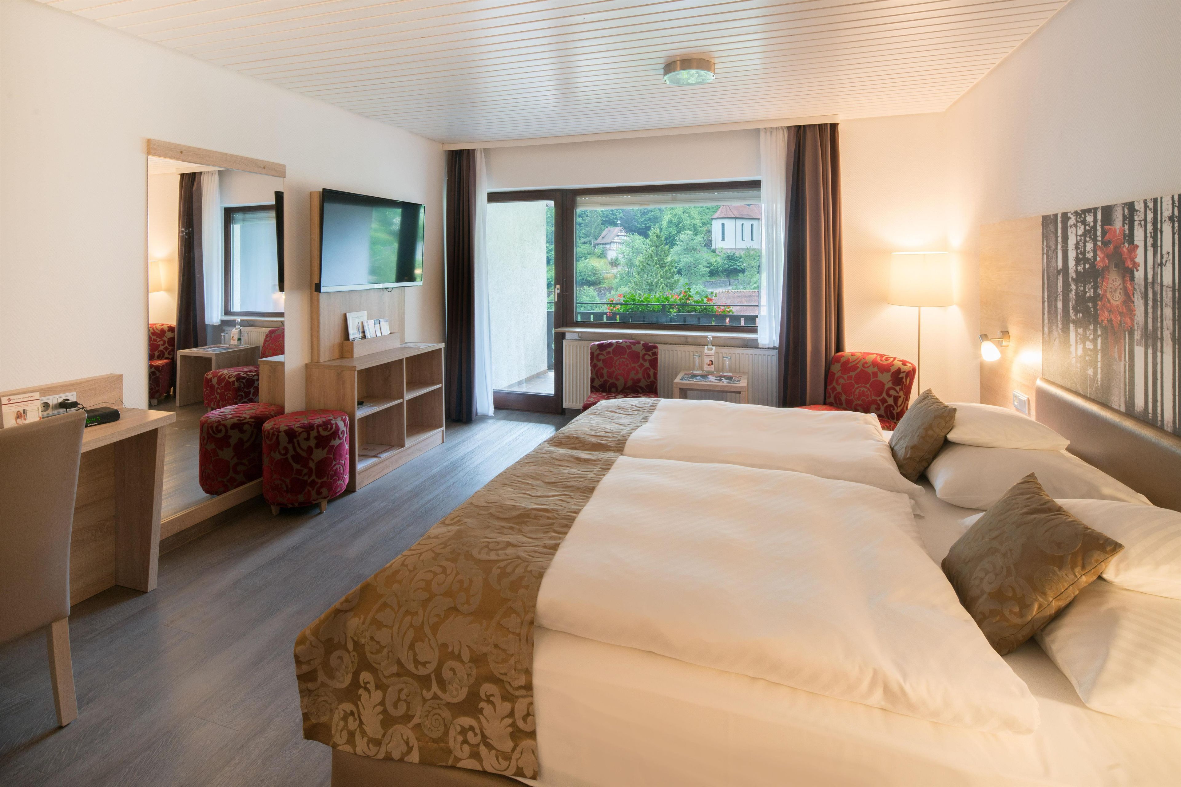 A hotel room with a large bed, a desk, a TV, and a window with a view of greenery.