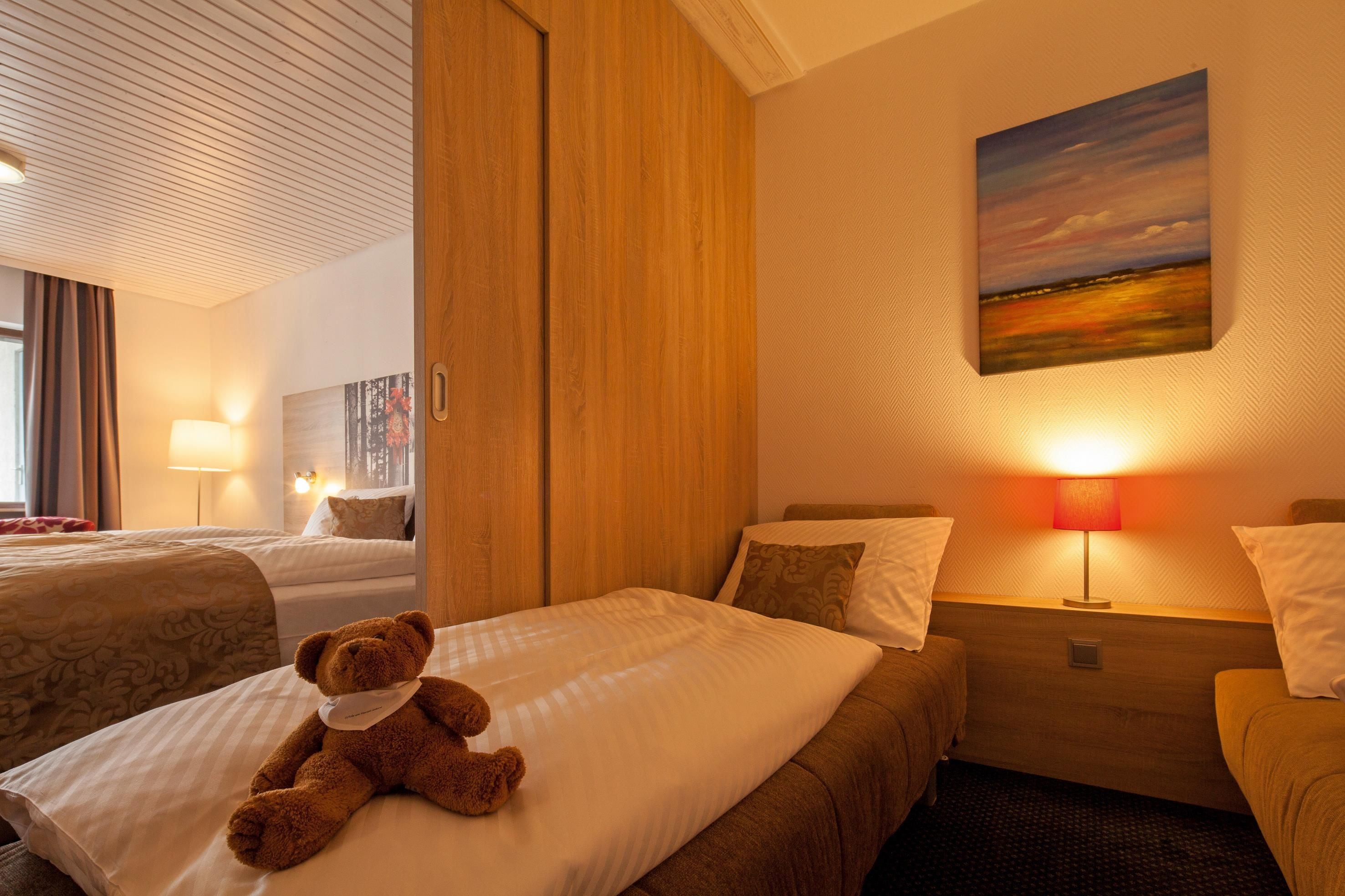 A hotel room with two beds, a teddy bear on one bed, a painting on the wall, and a lamp on the nightstand.
