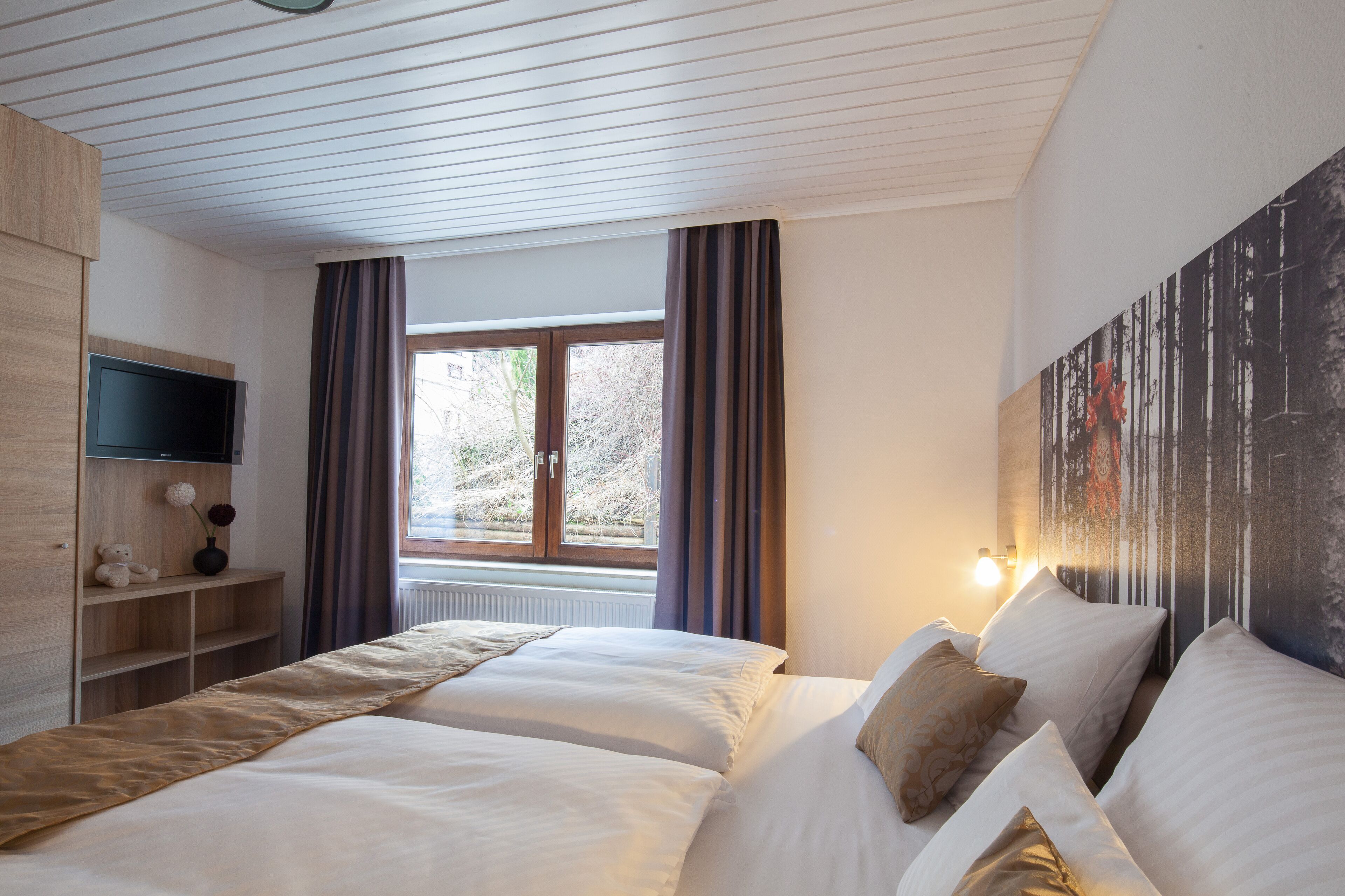 Et hotelværelse med to senge, et fjernsyn ophængt på væggen, et vindue med gardiner og et maleri på væggen.