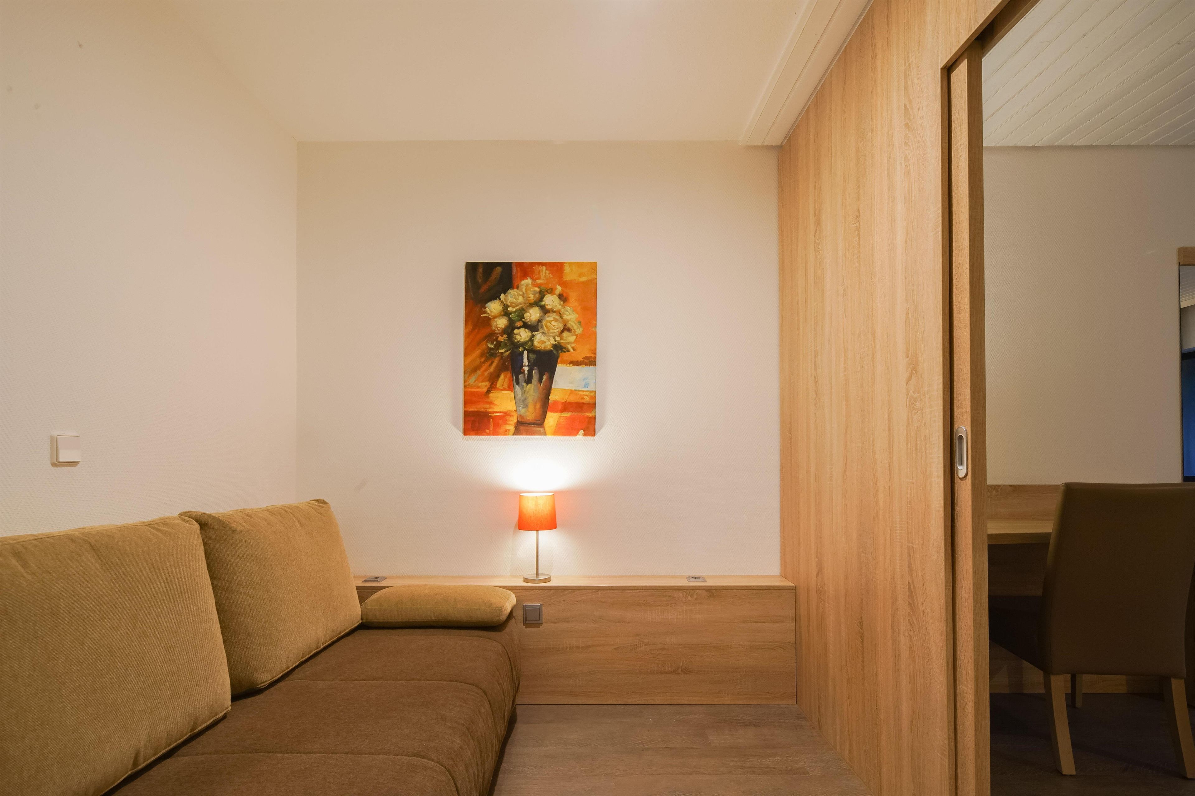 A room with a brown sofa, a table lamp, and a painting of flowers on the wall.