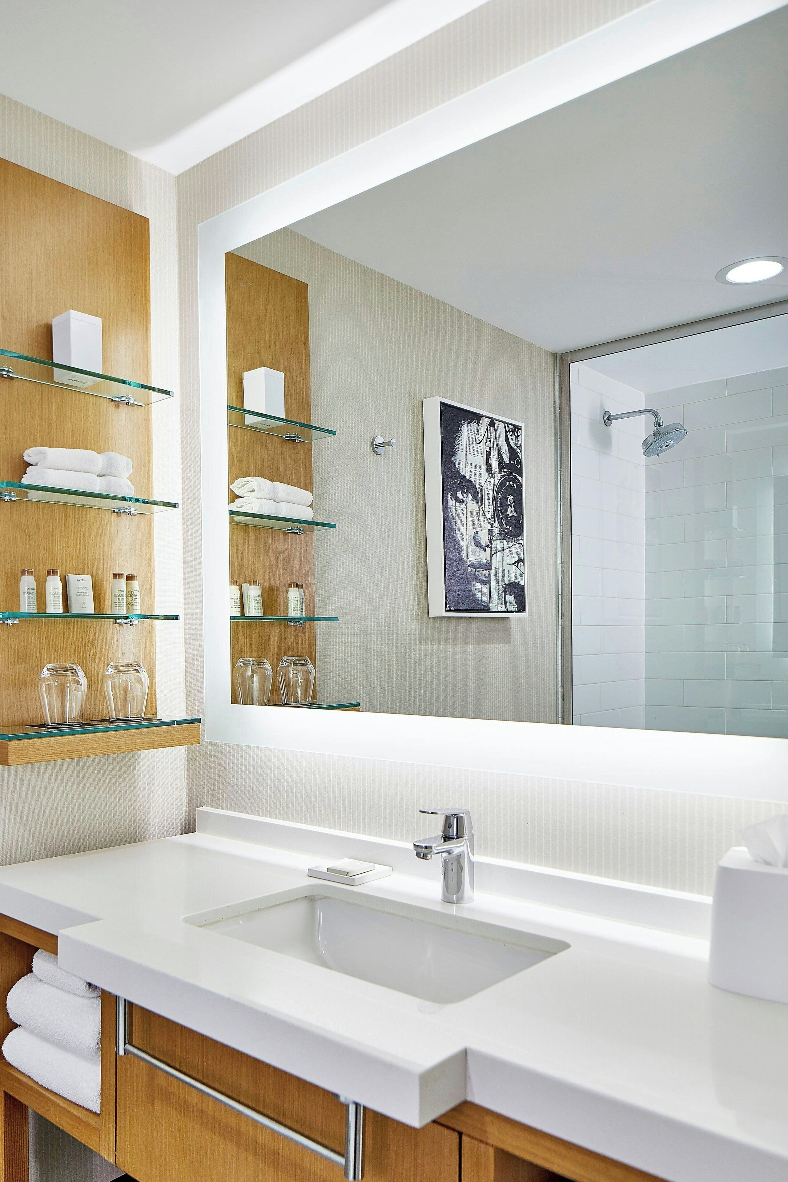 A modern bathroom with a large mirror, glass shelves with toiletries, a white sink, and a shower area.