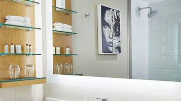 A modern bathroom with a large mirror, glass shelves with toiletries, a white sink, and a shower area.