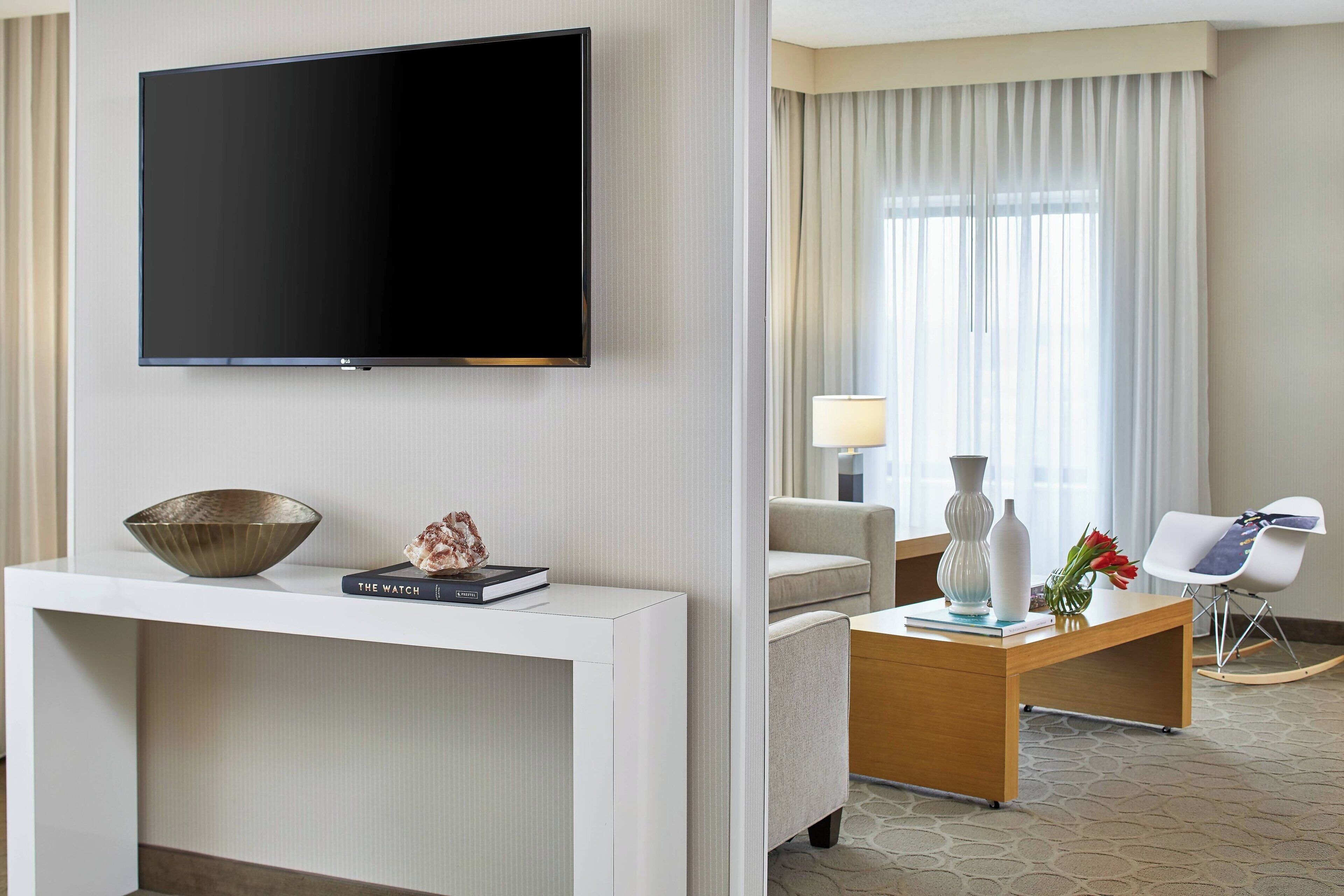 A modern hotel room with a flat-screen TV mounted on the wall, a white console table with a bowl and a book, a wooden coffee table with vases and flowers, a beige sofa, and a white chair.