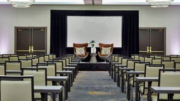 Une salle de conférence avec des rangées de chaises et de tables, un grand écran et deux fauteuils décoratifs ornés de vases.