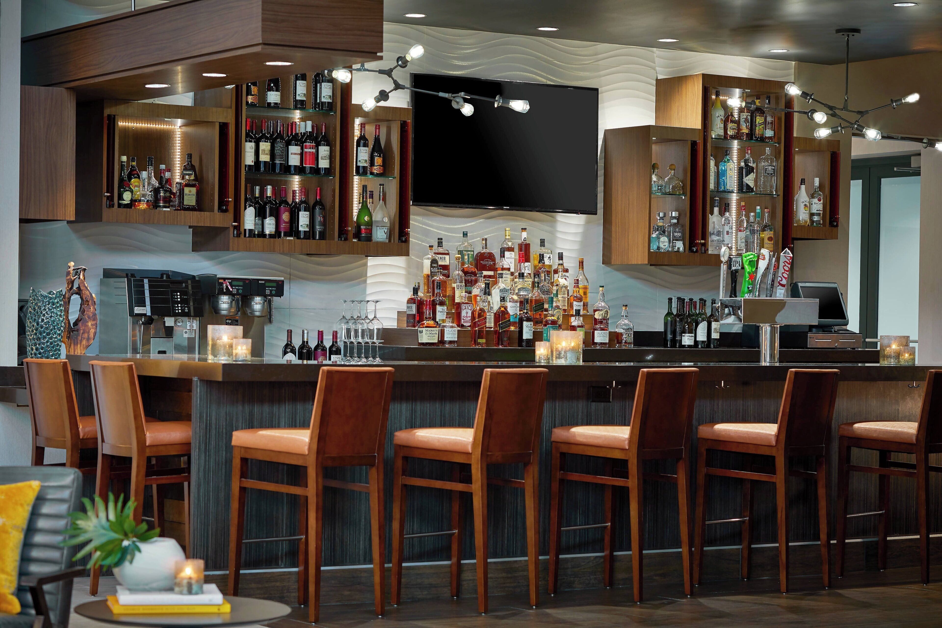 A bar area with multiple liquor bottles, a TV mounted on the wall, and bar stools.