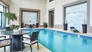 Ein Hallenbad mit Liegestühlen und einem Tisch, umgeben von Blumentöpfen und einem großen Fenster mit Blick.
