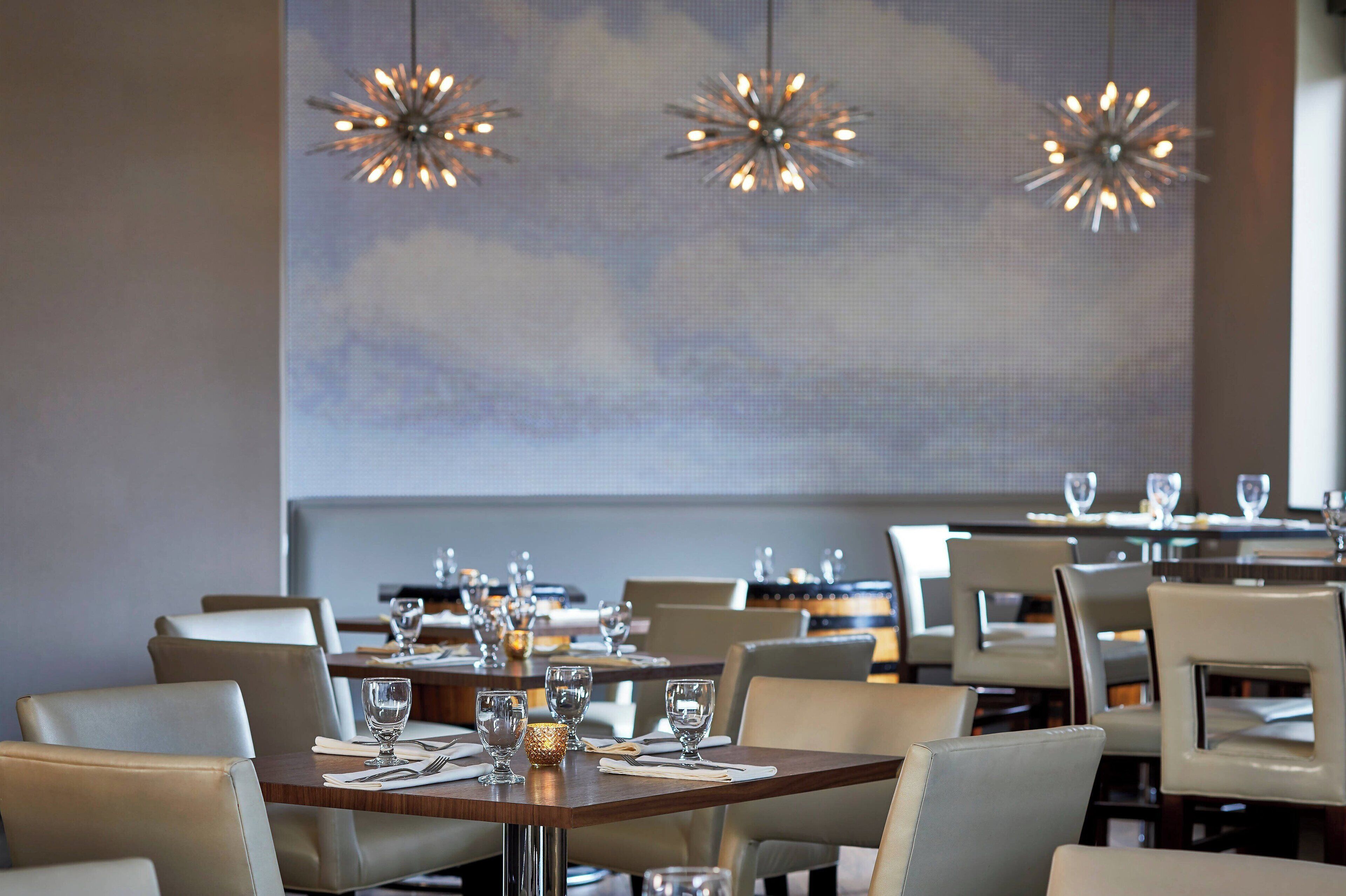 Restaurante com mesas preparadas para refeições, com lustres modernos e um grande mural na parede.