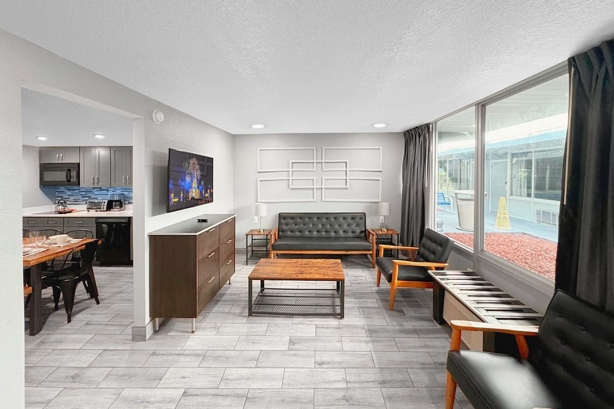Ein modernes Wohnzimmer mit einer Couch, einem Couchtisch und einem Fernseher. Es gibt eine Küchenzeile mit einer Mikrowelle und einen Esstisch mit Stühlen. Das Zimmer verfügt über große Fenster und einen Blick nach draußen.