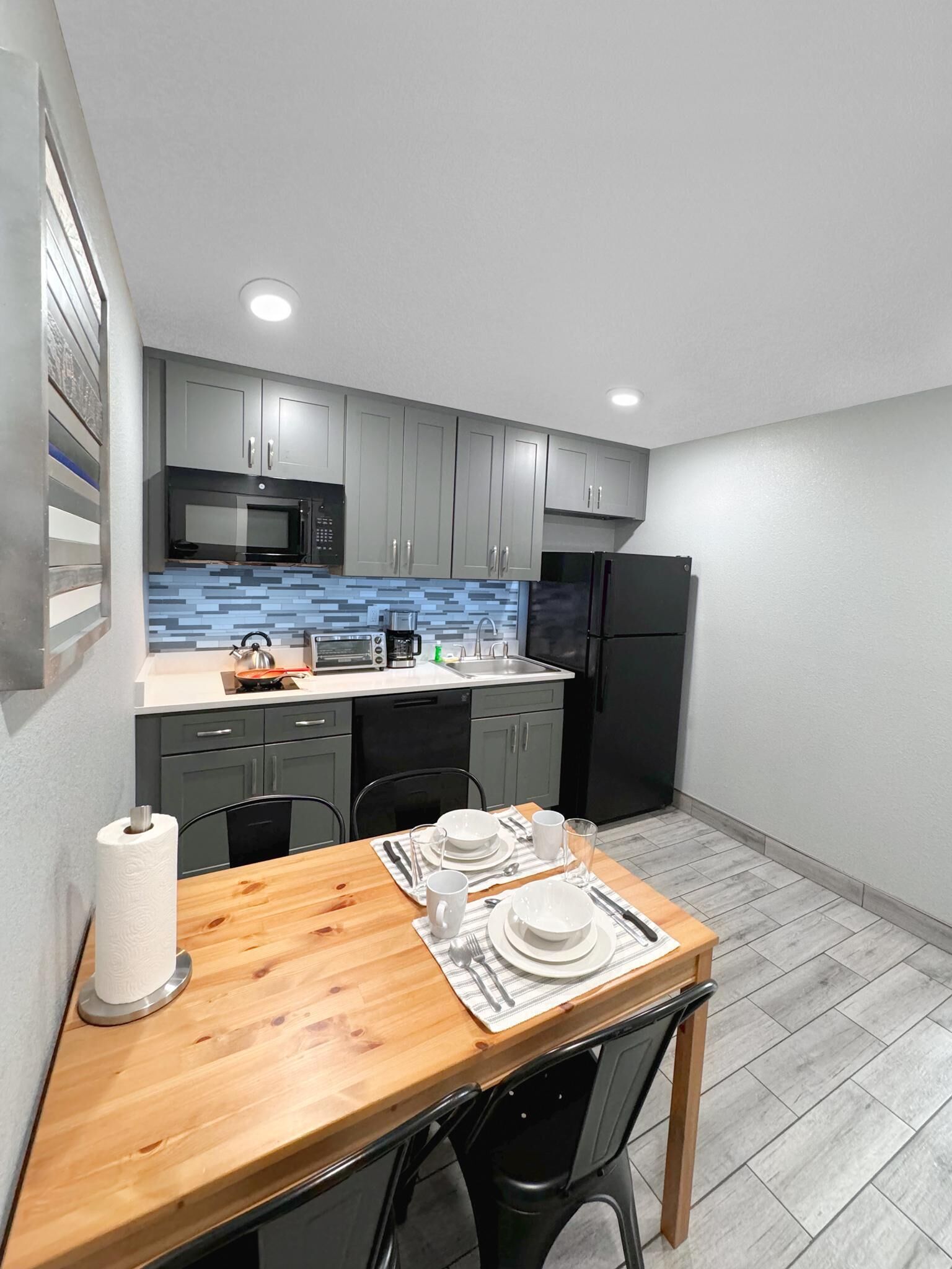 A compact kitchen with grey cabinets, a black refrigerator, and a wooden dining table set for two.