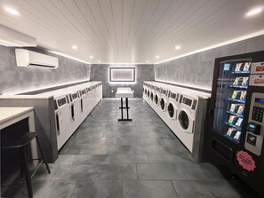 A modern laundry facility with multiple washing machines, a vending machine, and a central table. - Celebration Suites (Kissimmee)