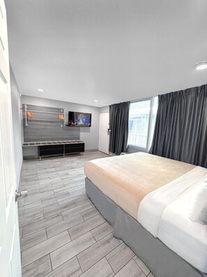 A modern hotel room with a bed, a TV mounted on the wall, a seating area with a bench, and a window with curtains. - Celebration Suites (Kissimmee)