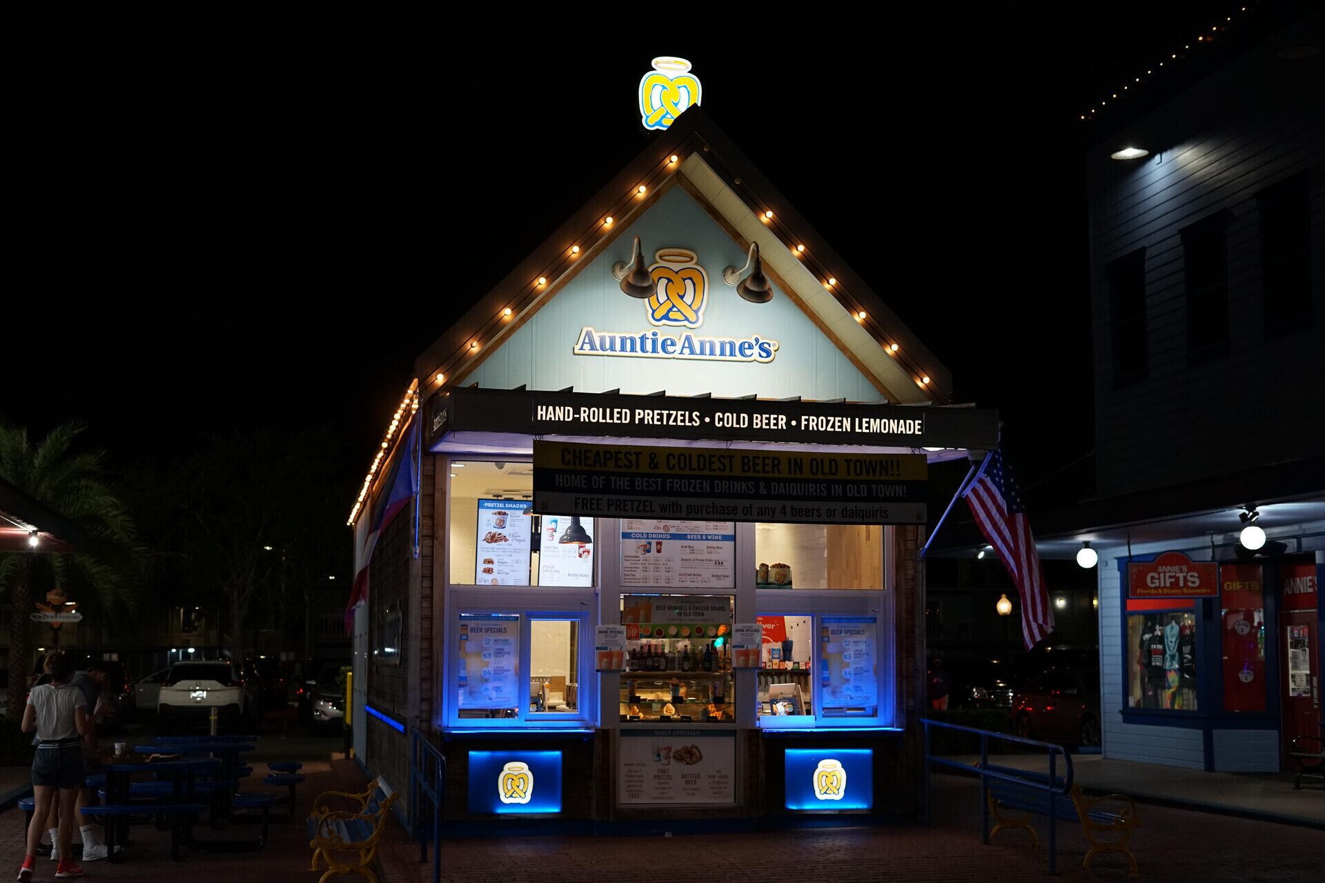 A small, brightly lit building with a sign reading 'AuntieAnne's' offering hand-rolled pretzels, cold beer, and frozen lemonade.