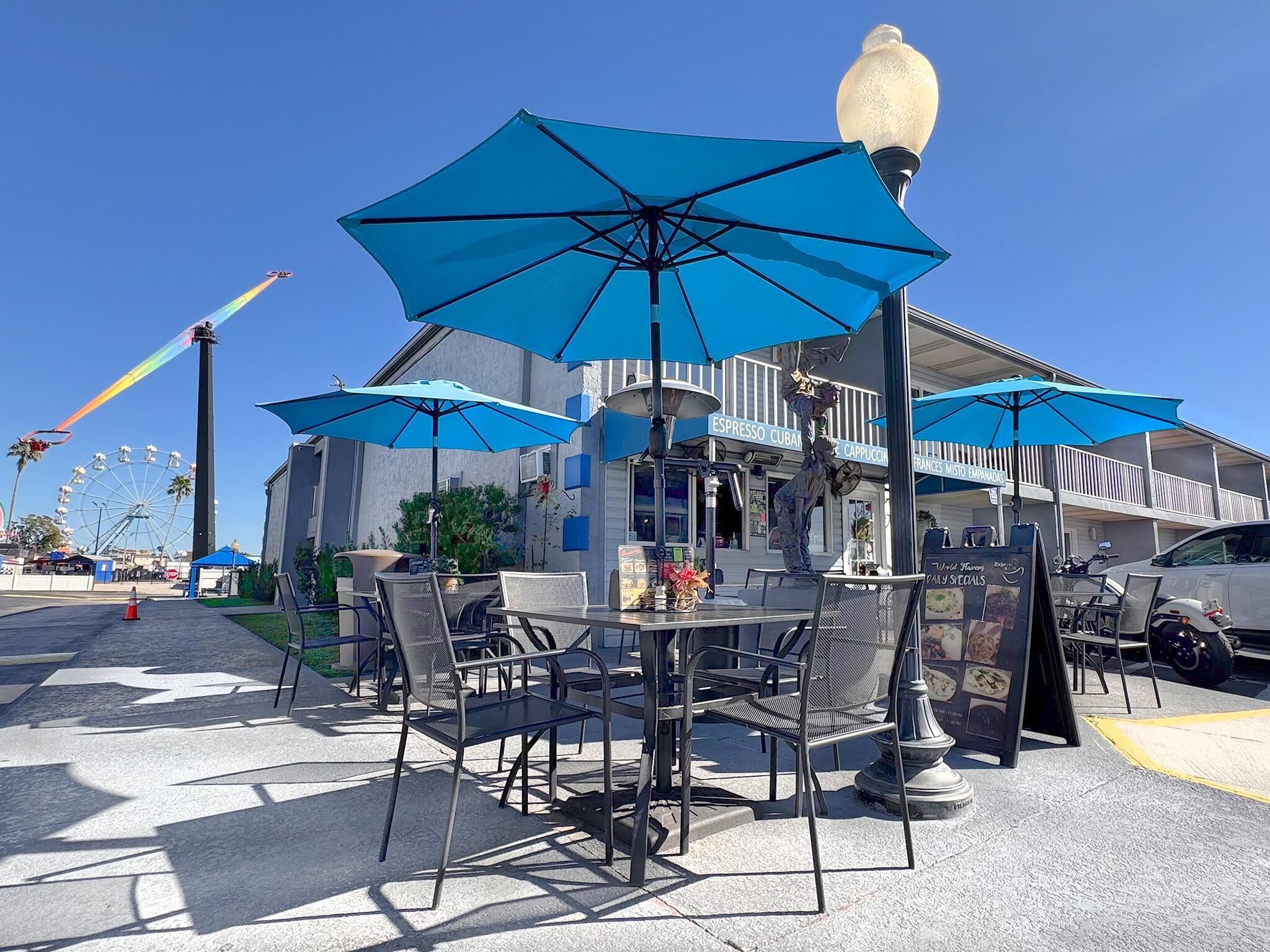 An outdoor seating area with tables and chairs under blue umbrellas, a street lamp, and a building with signage for a coffee shop.