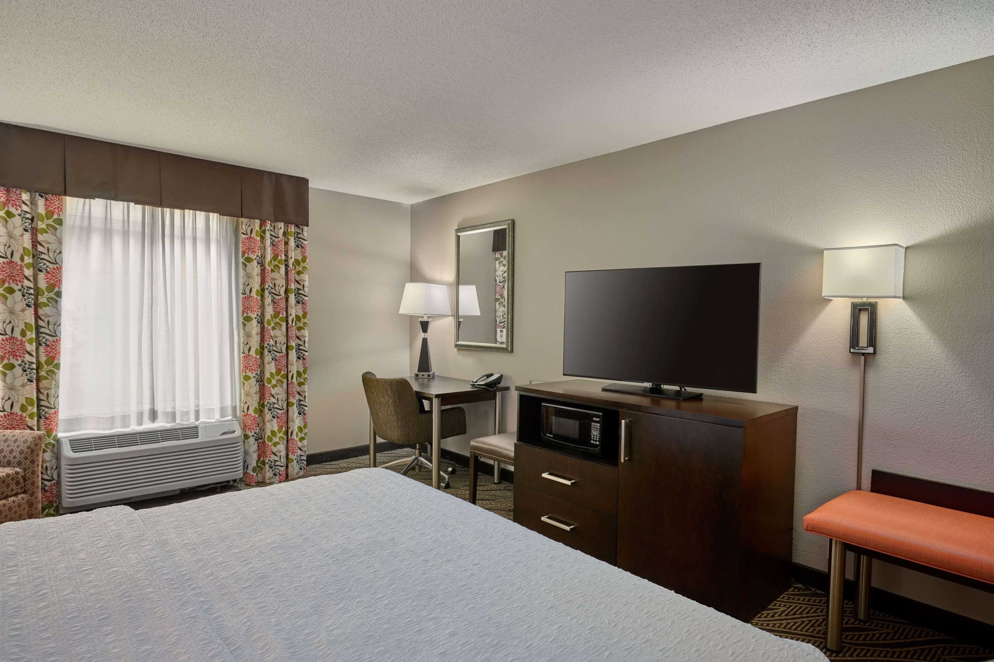 A hotel room with a bed, a desk with a chair, a television, a microwave, a lamp, and a bench.