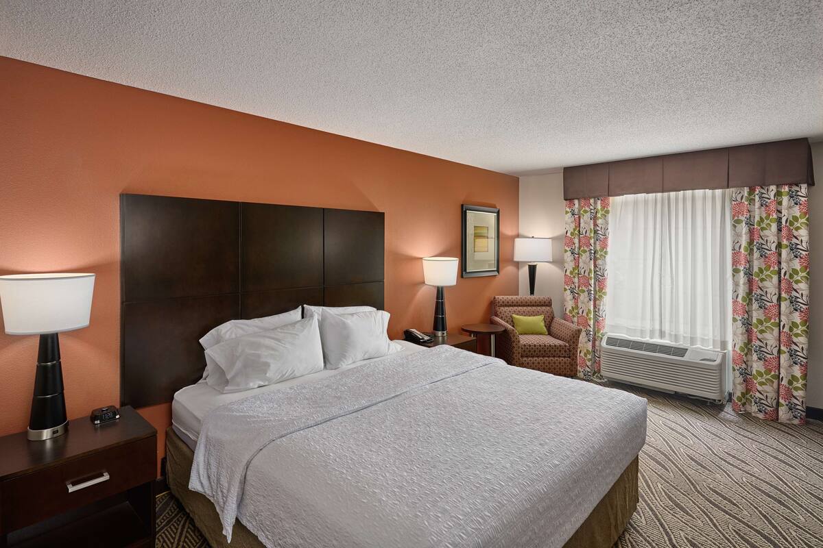 A hotel room with a large bed, two bedside tables with lamps, a chair, a window with floral curtains, and an air conditioning unit.
