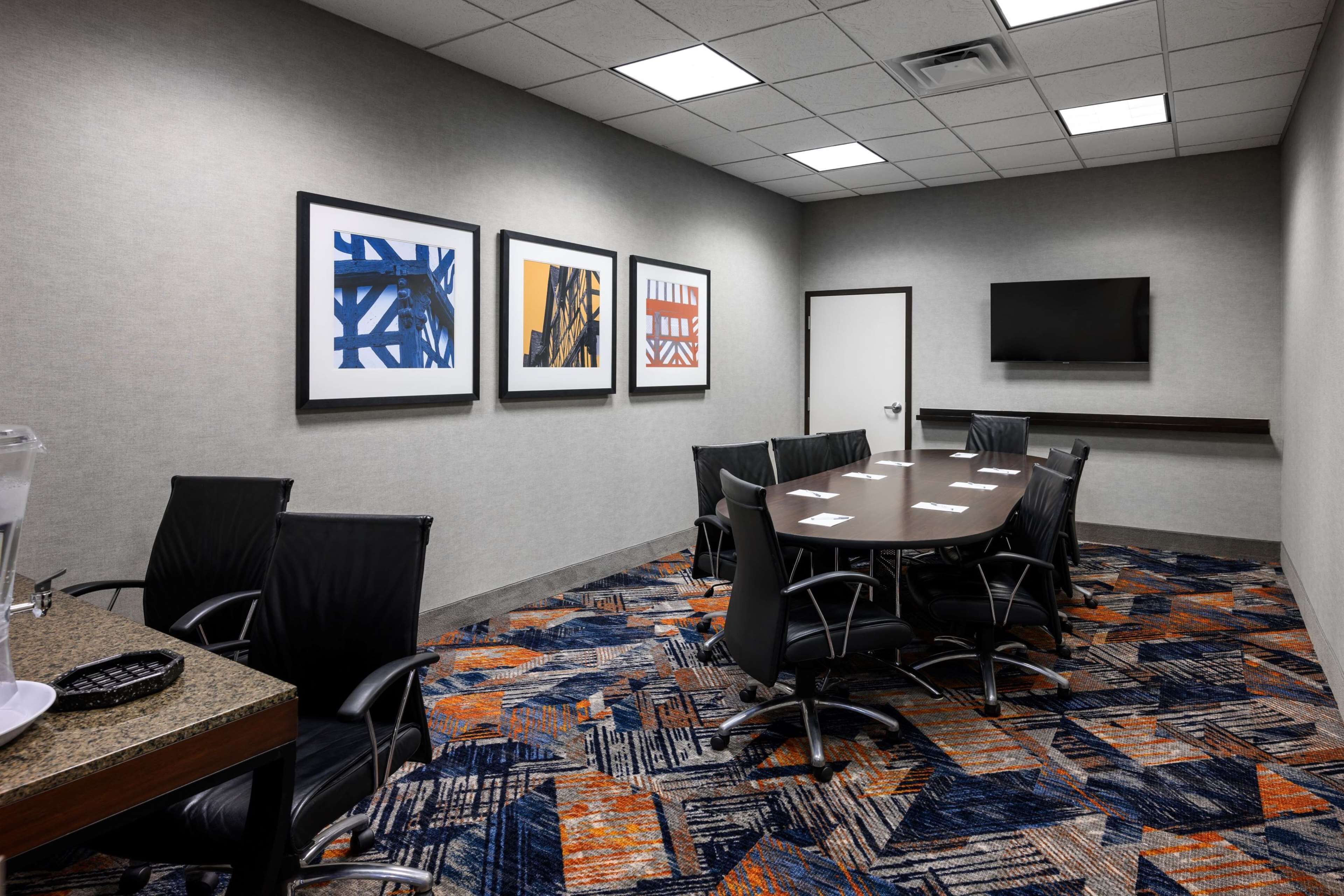A conference room with a round table, black chairs, framed artwork on the wall, a flat-screen TV, and a patterned carpet.