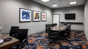 A conference room with a round table, black chairs, framed artwork on the wall, a flat-screen TV, and a patterned carpet.