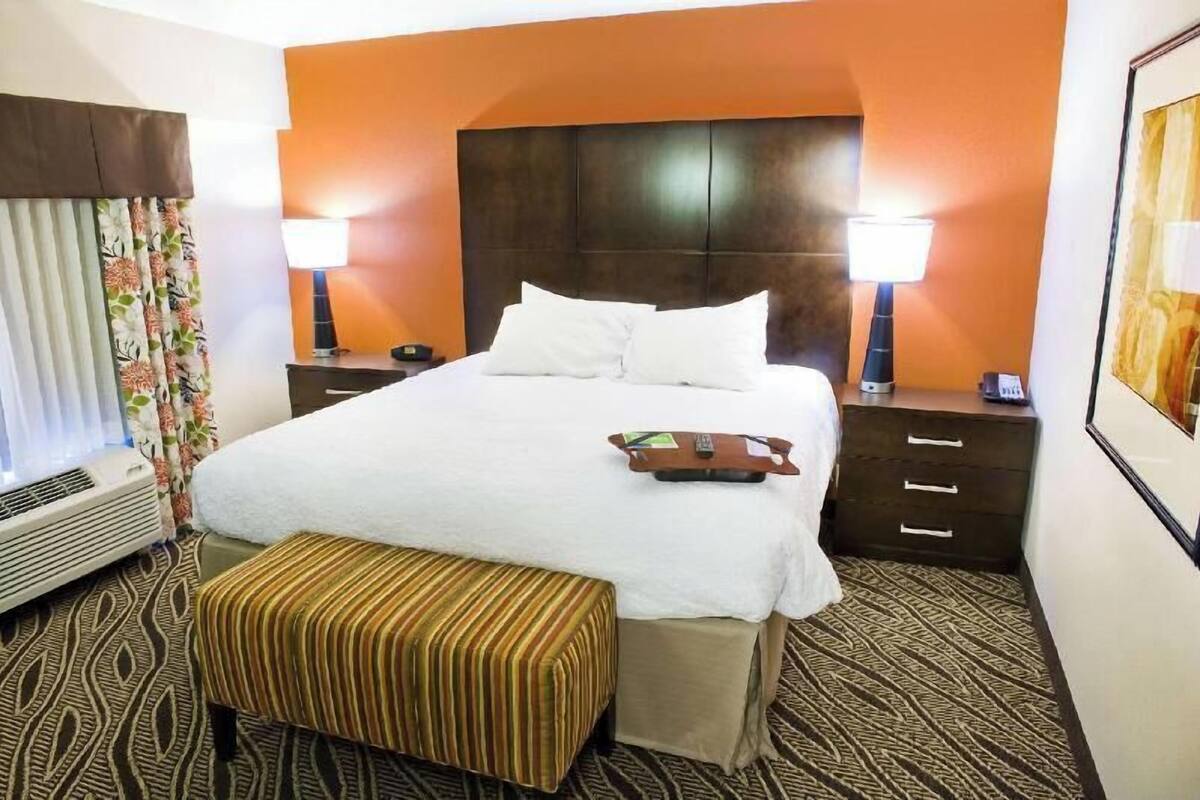 A hotel room with a large bed, two bedside tables, a bench, and a wall-mounted heater.