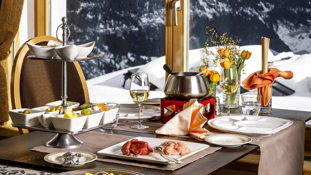 A dining table set with plates, bowls, and a wine glass, with a mountain view through a window. - Eiger Mürren Swiss Quality Hotel (Mürren)