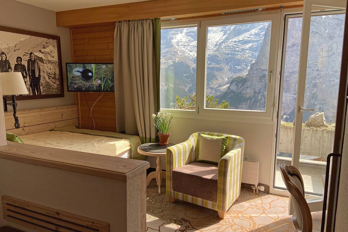 Een hotelkamer met een bed, een kleintje tafeltje met een bloempot, een gestreept fauteuil en een televisie aan de muur.