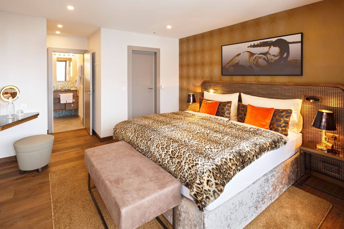 Een moderne slaapkamer met een groot bed, een bankje, een nachtkastje met lampen, en een badkamer zichtbaar door een geopende deur.