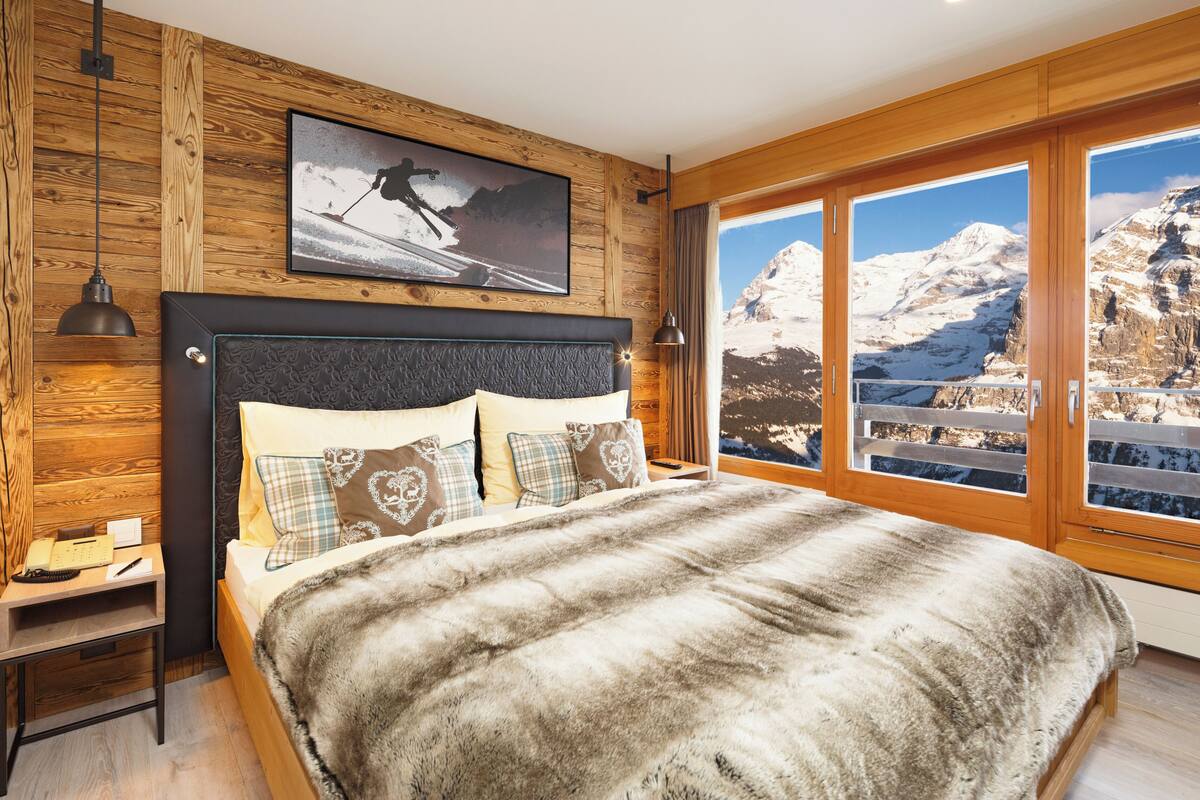 Een slaapkamer met een groot bed, houten wanden, een afbeelding met een winters thema en uitzicht op de bergen.