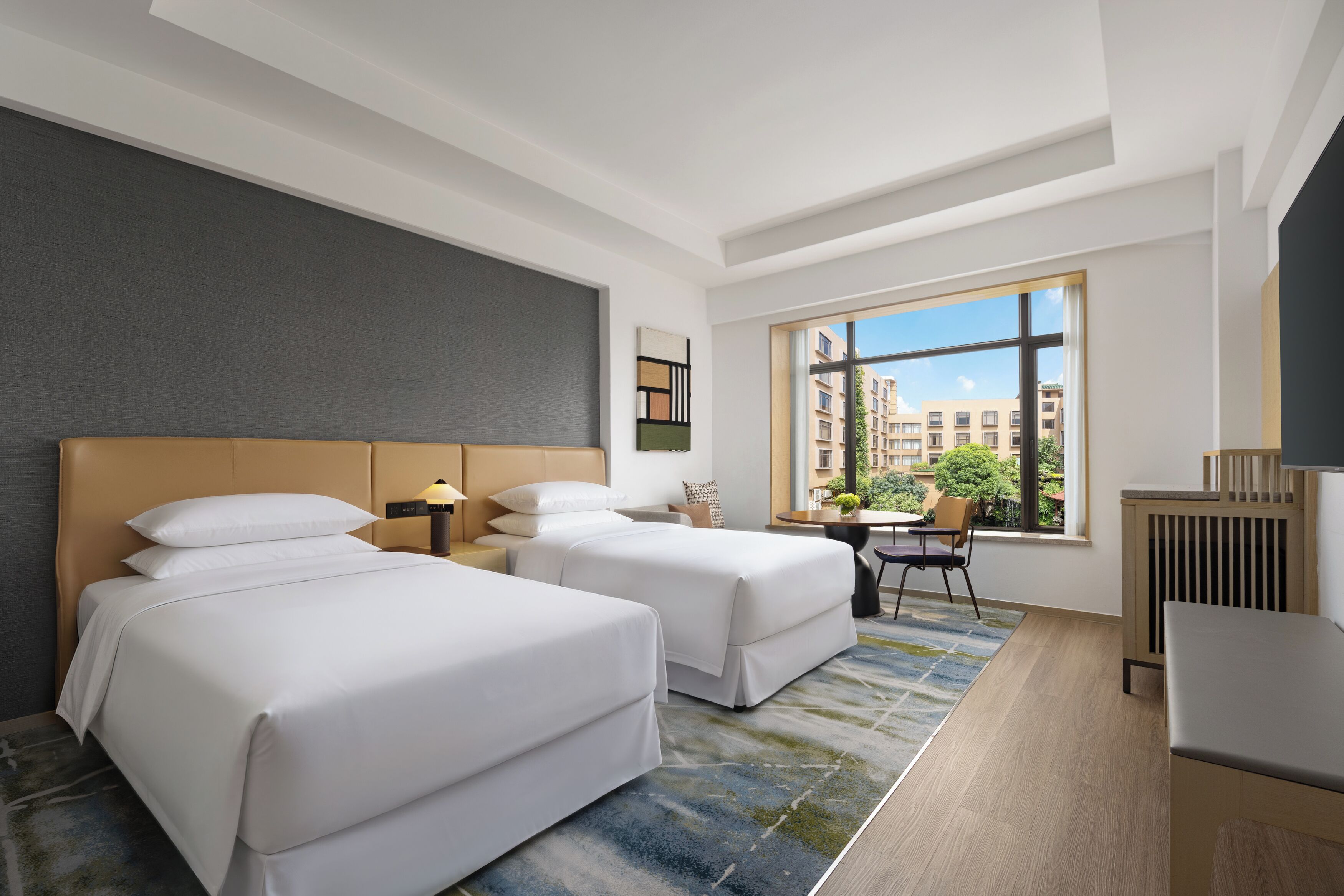 A hotel room with two beds, a desk, a chair, a radiator, and a window with a view of buildings.