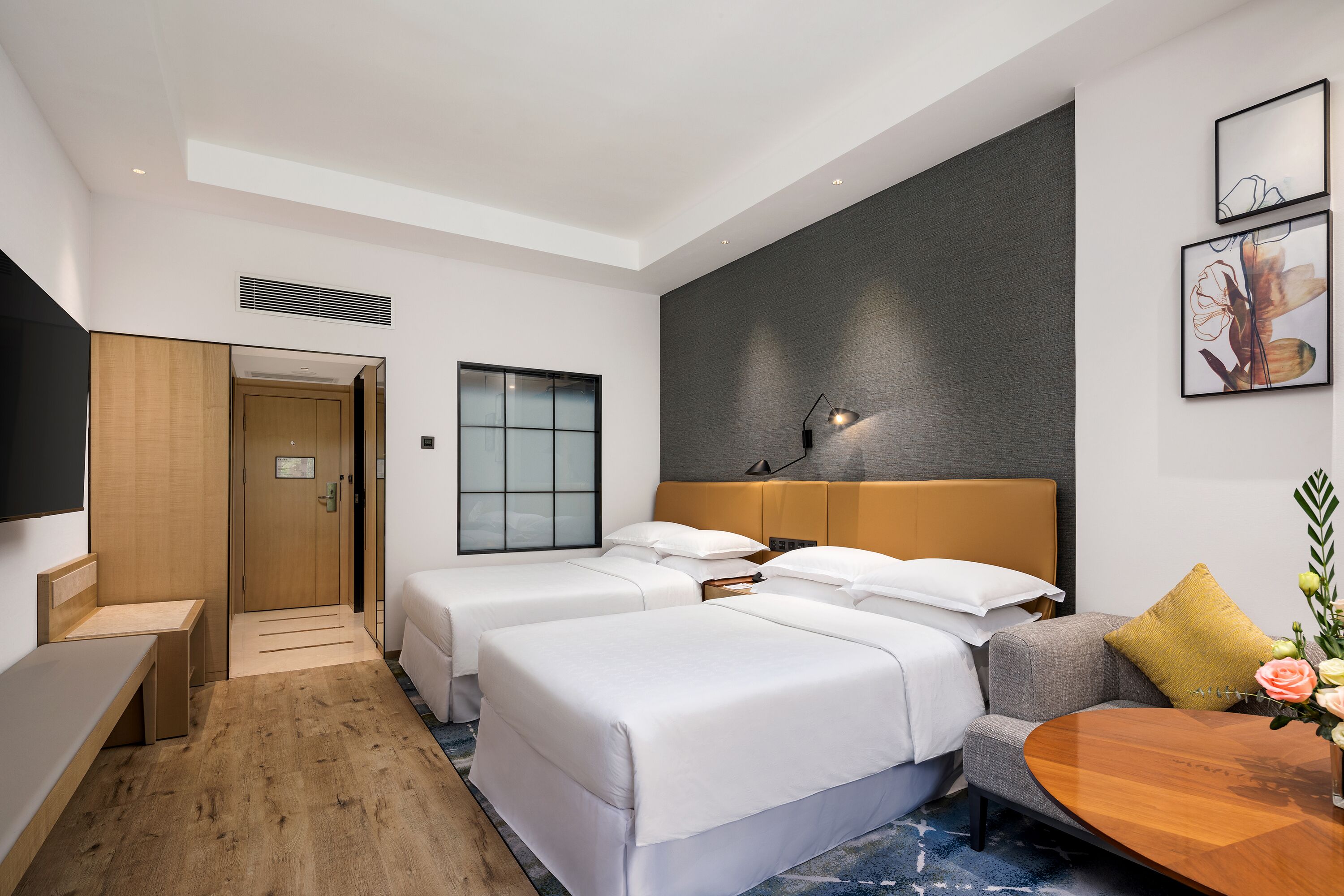 Habitación de hotel con dos camas individuales, un sofá, un banco de madera, una mesita y un televisor instalado en la pared.