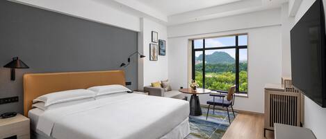 A modern hotel room with a large bed, a desk, a chair, a TV, and a window with a view of greenery.