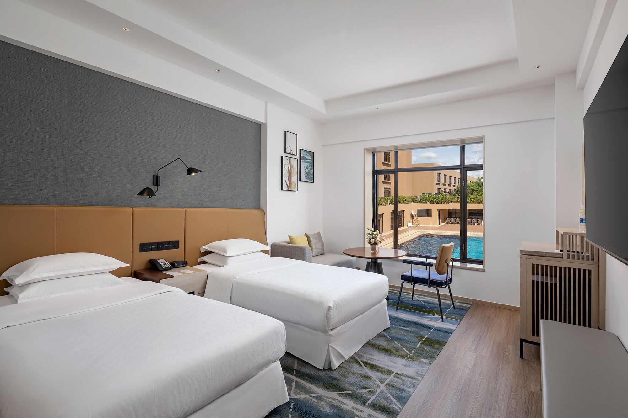 A hotel room with two beds, a desk, a chair, a small table, a large window with a view of a pool and building, and a TV.