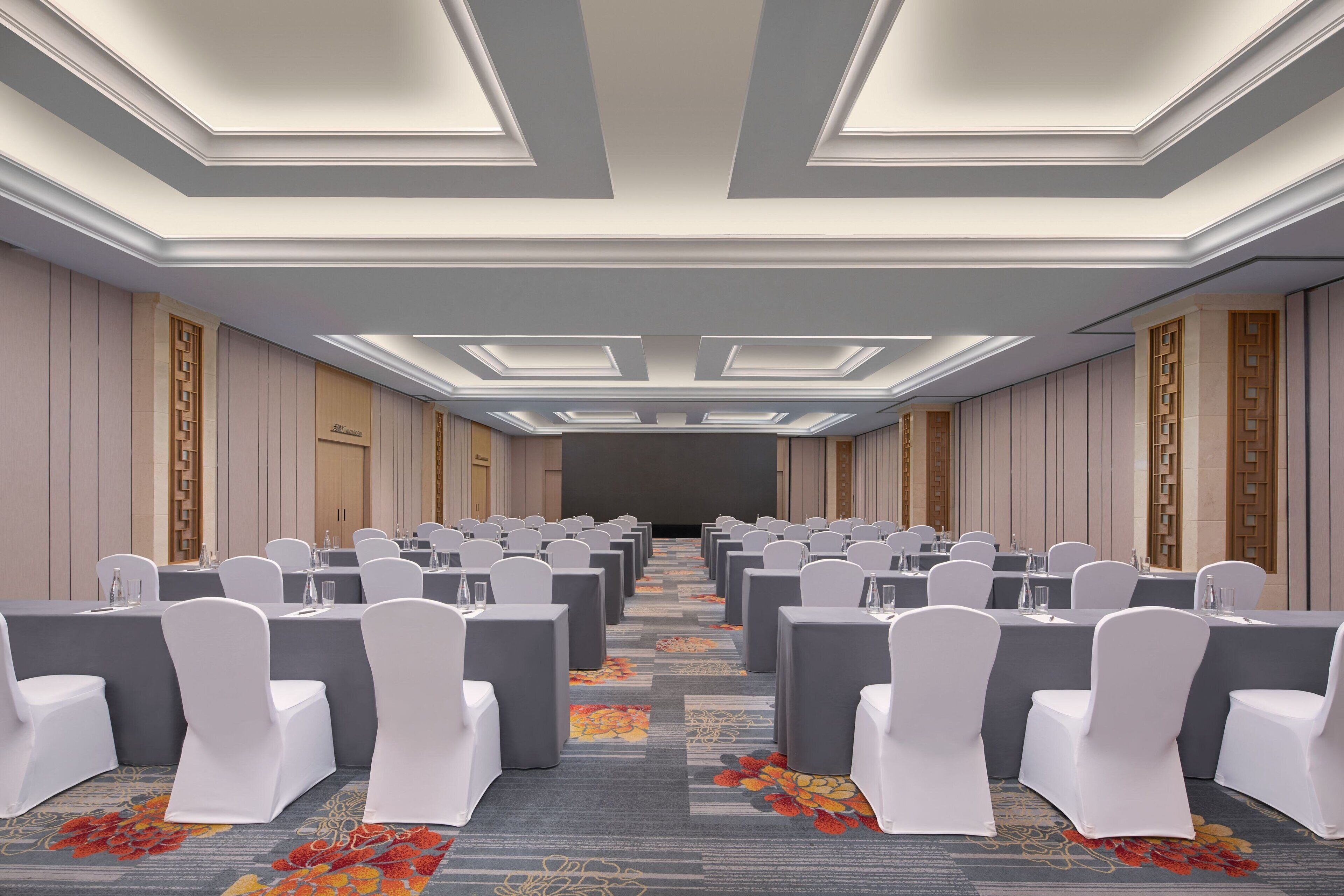 A conference room with rows of tables and chairs, a carpeted floor, and a modern ceiling design.