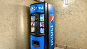 A Pepsi vending machine in a corner with a textured wall and a small window. - Quality Inn San Angelo (San Angelo)
