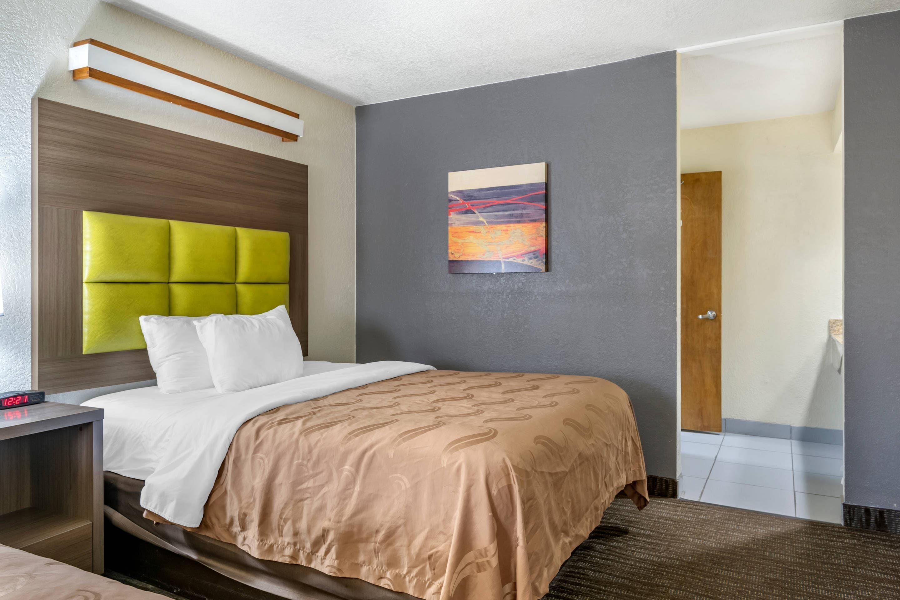 A hotel room with a bed, a nightstand, a wall-mounted painting, and a door leading to another room.