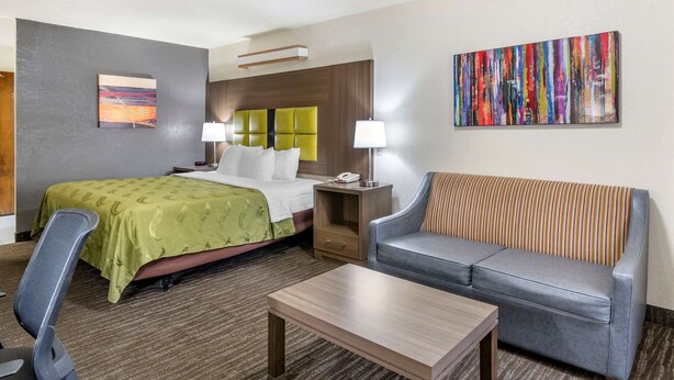 A hotel room with a bed, a grey sofa, a wooden coffee table, a desk, and two lamps. - Quality Inn San Angelo (San Angelo)
