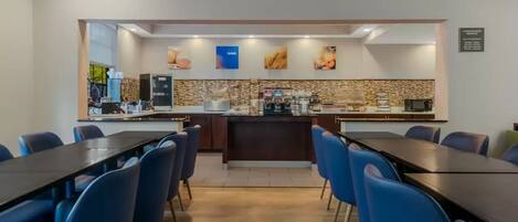 A modern hotel dining area with a central bar, blue chairs, and a tiled backsplash.